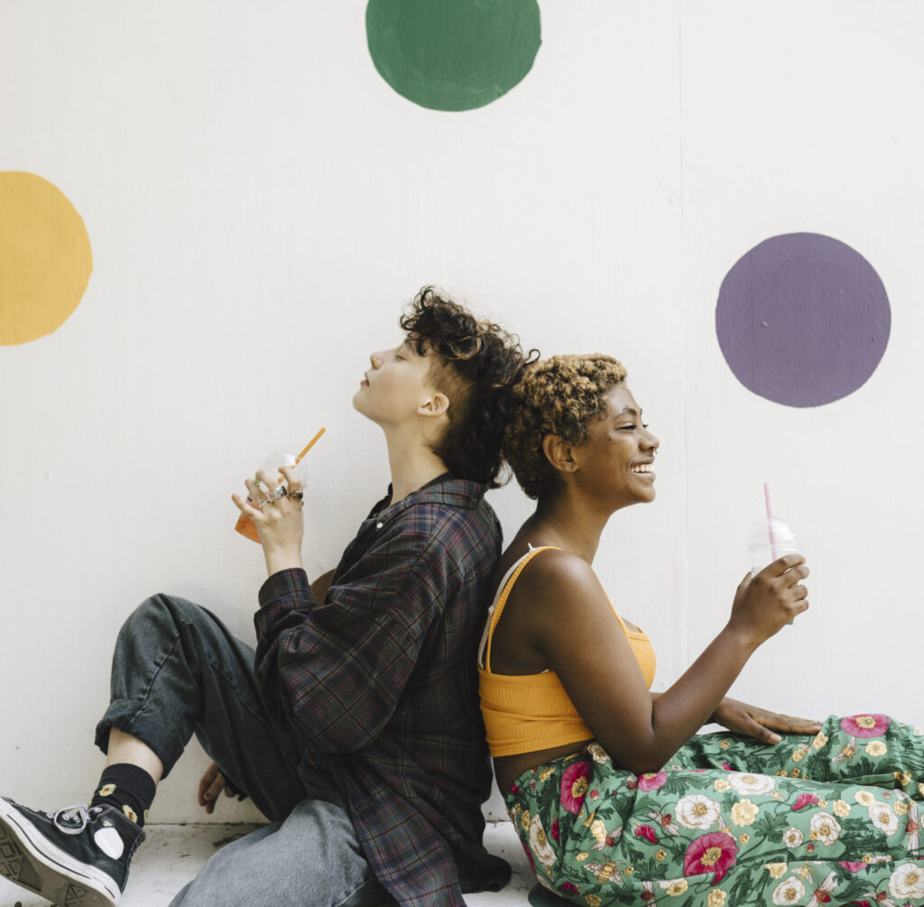 Teenage boy sitting back-to-back with his smiling non-binary teenage friend in front of a colorful wall